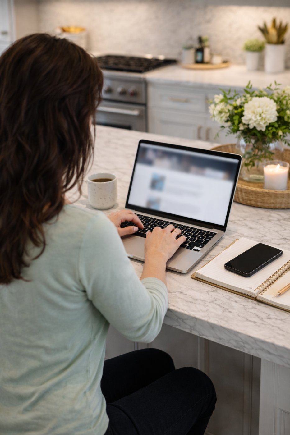 A woman working at her laptop in a bright kitchen with coffee, flowers and a notebook