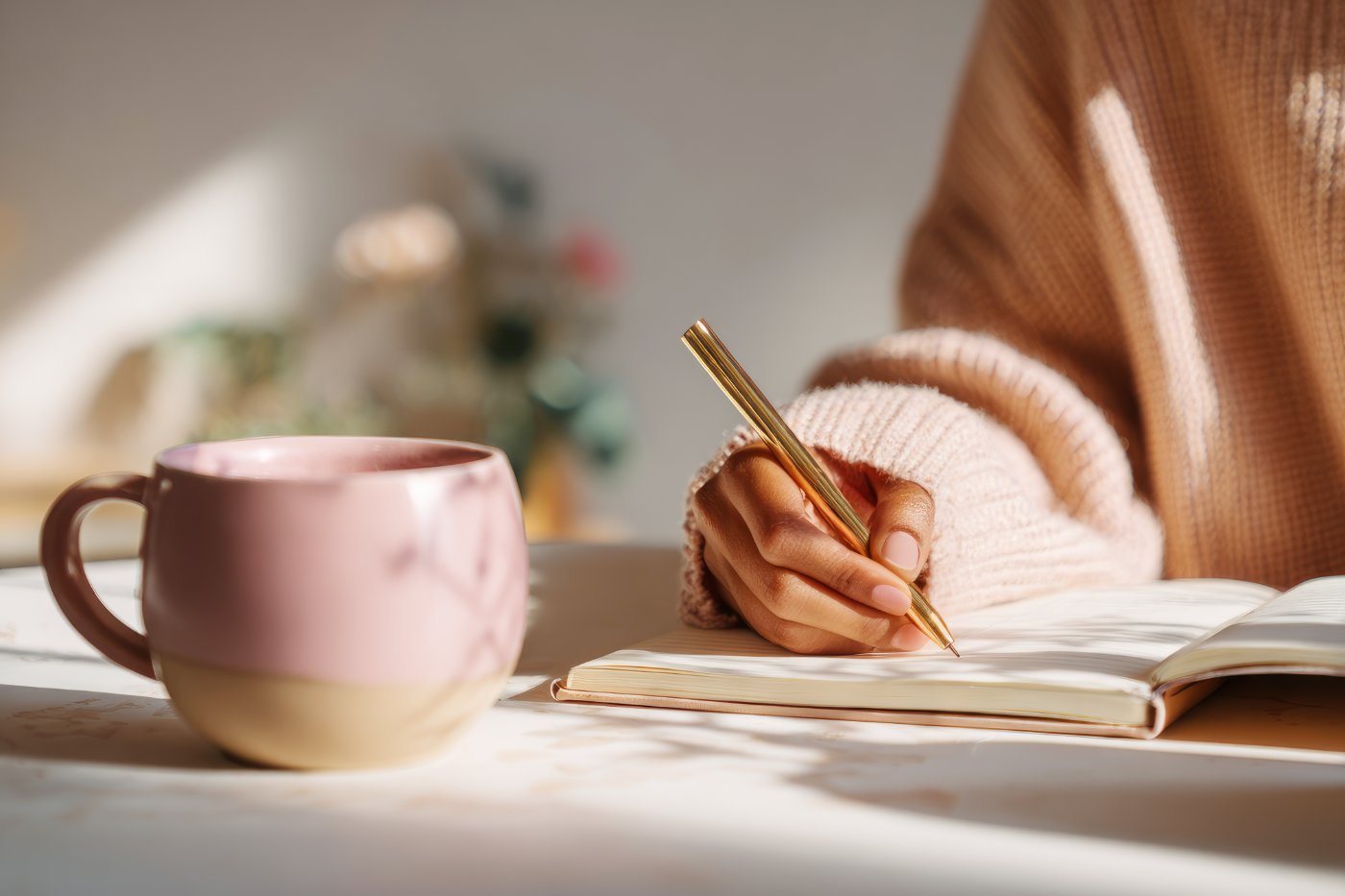 A woman journaling with a gold pen beside a pink cup of tea in soft daylight